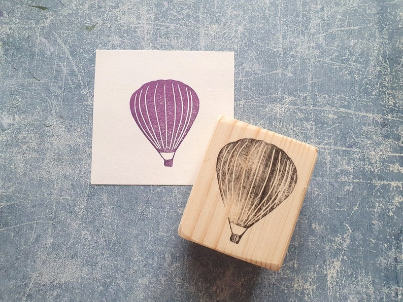 Pode incluir: Um carimbo de bloco de madeira com um desenho de bal&atilde;o de ar quente roxo. O carimbo est&aacute; em um cart&atilde;o branco com um desenho de bal&atilde;o de ar quente roxo.