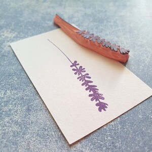 May include: A wooden stamp with a purple lavender design. The stamp is on a white card and has been used to create an impression of the lavender.
