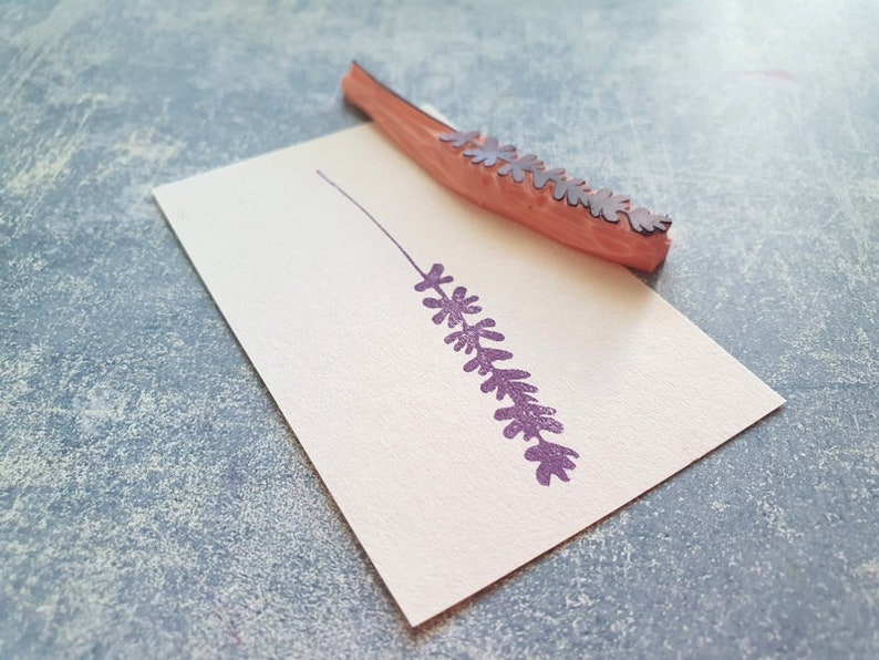 May include: A wooden stamp with a purple lavender design. The stamp is on a white card and has been used to create an impression of the lavender.