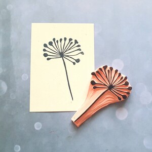 May include: A wooden stamp with a black silhouette of a dandelion flower. The stamp is orange and has a textured surface. The stamp is next to a printed image of the dandelion on a yellow card.
