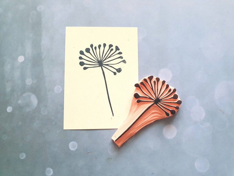 May include: A wooden stamp with a black silhouette of a dandelion flower. The stamp is orange and has a textured surface. The stamp is next to a printed image of the dandelion on a yellow card.