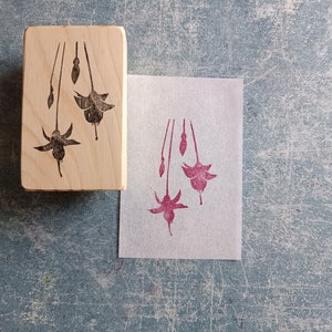 May include: A wooden block stamp with a design of three fuchsia flowers. The stamp is used to create a pink impression on a piece of paper.