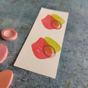 May include: Two pink and green abstract shapes printed on a white card. The shapes are overlapping and have a rough texture.