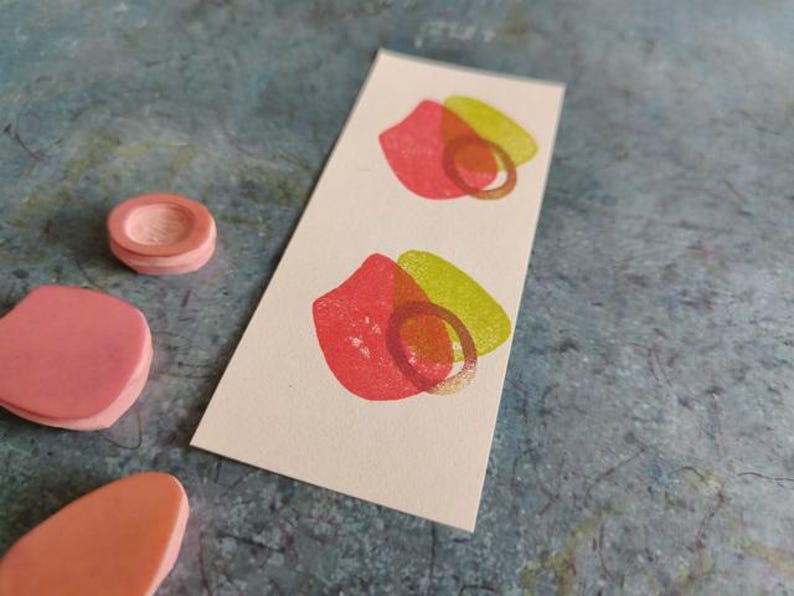 May include: Two pink and green abstract shapes printed on a white card. The shapes are overlapping and have a rough texture.