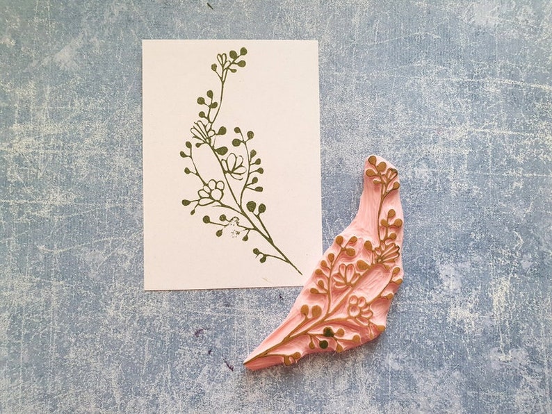 May include: A pink rubber stamp with a gold floral design. The stamp is shaped like a bird with its wings spread out. The design is similar to the one on the white card next to it.