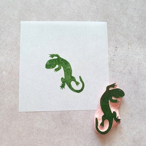 May include: A green lizard stamp and its impression on white paper. The stamp is made of pink rubber with a detailed lizard design. The stamped image is a vibrant green, showcasing the lizard's form.