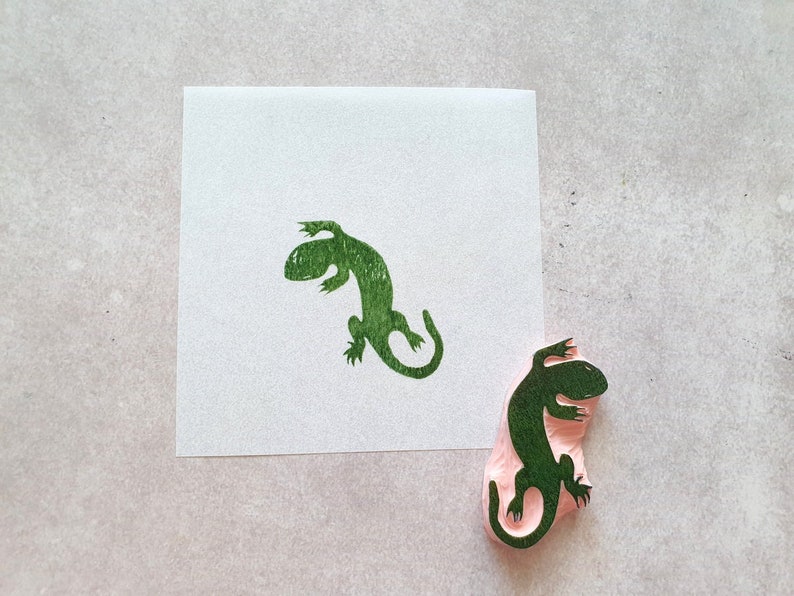 May include: A green lizard stamp and its impression on white paper. The stamp is made of pink rubber with a detailed lizard design. The stamped image is a vibrant green, showcasing the lizard's form.