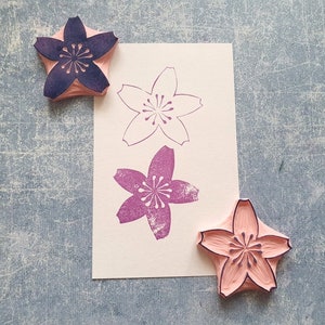 Peut inclure: Trois tampons en bois avec un motif de fleurs de cerisier. Les tampons sont dans des tons de rose et de violet. Les tampons sont sur une surface textur&eacute;e grise.