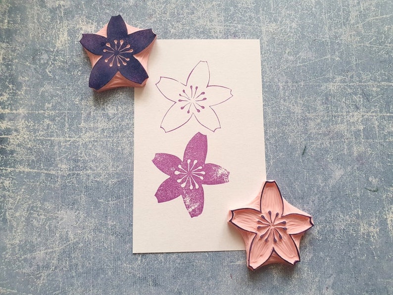 Peut inclure: Trois tampons en bois avec un motif de fleurs de cerisier. Les tampons sont dans des tons de rose et de violet. Les tampons sont sur une surface textur&eacute;e grise.