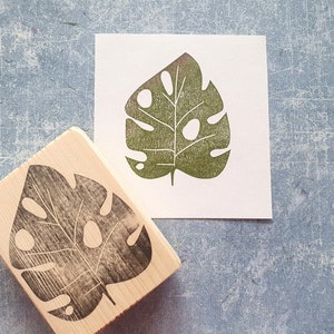 May include: A wooden stamp with a leaf design and a printed image of the same leaf. The leaf is green with several holes and a stem. The stamp is rectangular with the leaf design carved into the bottom.