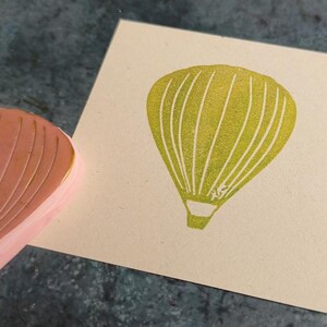 Pode incluir: Um close-up de um carimbo de borracha amarelo com um desenho de bal&atilde;o de ar quente. O carimbo est&aacute; em uma superf&iacute;cie bege.
