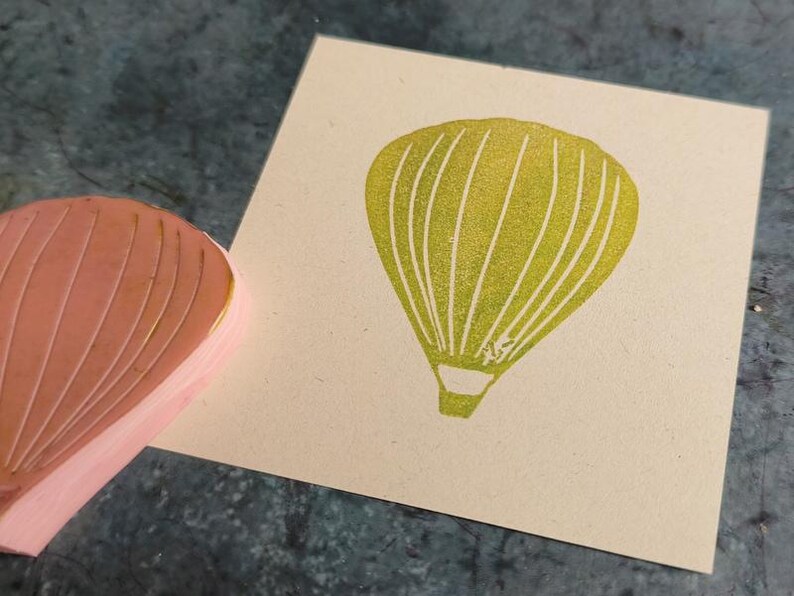 Pode incluir: Um close-up de um carimbo de borracha amarelo com um desenho de bal&atilde;o de ar quente. O carimbo est&aacute; em uma superf&iacute;cie bege.