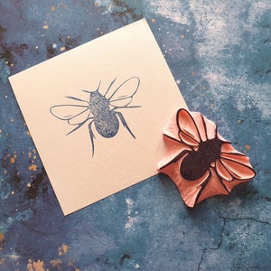 May include: A bee-shaped rubber stamp and a stamped image of a bee on a square piece of light tan paper. The stamp is pink with a black bee design. The paper is on a blue and gold speckled surface.