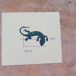 May include: A teal-colored lizard silhouette on a cream-colored square. The lizard is approximately 1.4 inches long and 2 inches tall. The image includes handwritten measurements in centimeters. The background is a textured, mottled surface.