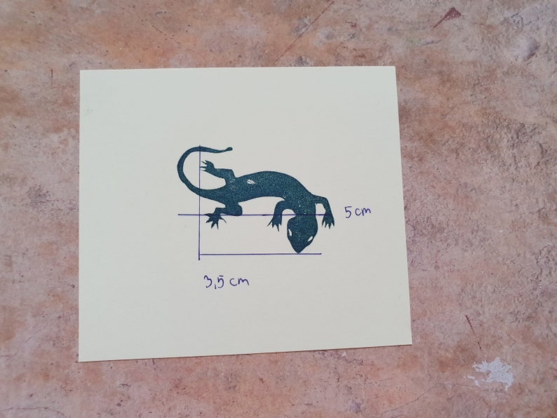 May include: A teal-colored lizard silhouette on a cream-colored square. The lizard is approximately 1.4 inches long and 2 inches tall. The image includes handwritten measurements in centimeters. The background is a textured, mottled surface.
