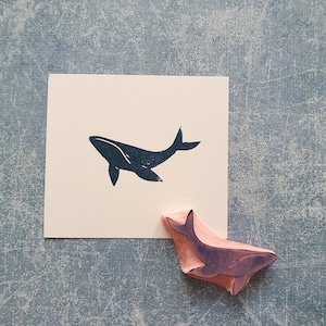 May include: A black whale silhouette rubber stamp with a wooden handle. The stamp is on a light gray surface with a white card showing the whale silhouette.