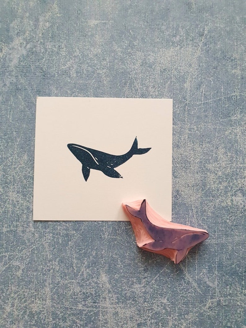 May include: A black whale silhouette rubber stamp with a wooden handle. The stamp is on a light gray surface with a white card showing the whale silhouette.