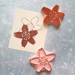 Peut inclure: Trois tampons en bois avec un motif de fleur de cerisier. Deux des tampons sont roses et un est brun.