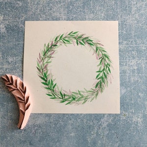Könnte beinhalten: Eine weiße Karte mit einem grünen und rosafarbenen Aquarell-Kranz-Design. Ein Holz-Stempel mit einem Blatt-Design befindet sich in der linken unteren Ecke des Bildes.