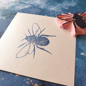 May include: A blue insect stamp impression on a cream-colored square of paper. A pink and black rubber stamp of the same insect is next to the impression. The background is a textured blue surface with gold flecks.