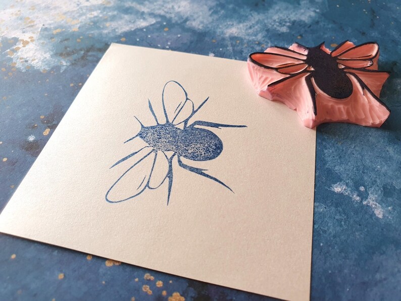 May include: A blue insect stamp impression on a cream-colored square of paper. A pink and black rubber stamp of the same insect is next to the impression. The background is a textured blue surface with gold flecks.
