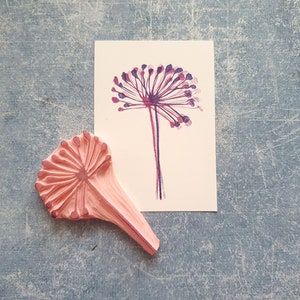 May include: A pink rubber stamp with a detailed design of a flower with a long stem. The stamp is next to a white card with a purple and pink watercolor illustration of a flower.