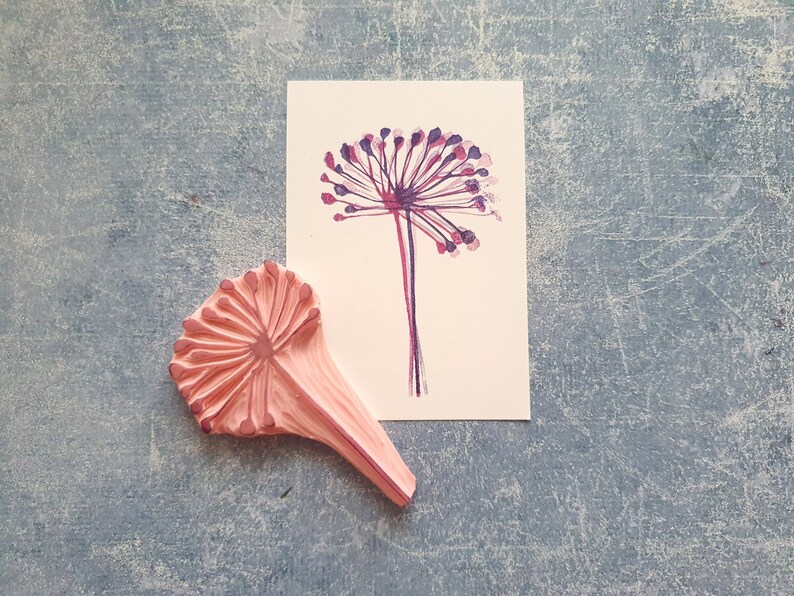 May include: A pink rubber stamp with a detailed design of a flower with a long stem. The stamp is next to a white card with a purple and pink watercolor illustration of a flower.