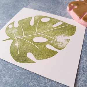 May include: A green monstera leaf print on white paper. The leaf design is stamped with a textured effect. A pink rubber stamp of a monstera leaf is visible in the background. The image is on a blue surface.