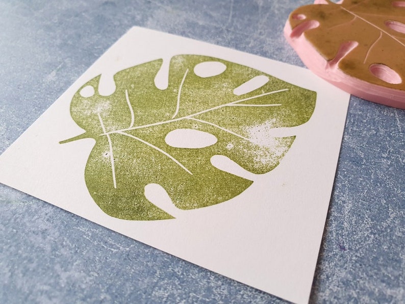 May include: A green monstera leaf print on white paper. The leaf design is stamped with a textured effect. A pink rubber stamp of a monstera leaf is visible in the background. The image is on a blue surface.
