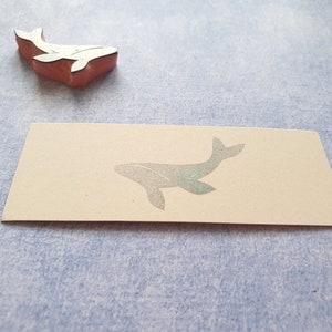 May include: A close-up of a blank card with a whale silhouette printed on it. The whale is a light gray color and is facing to the right. The card is a light beige color and has a slightly textured surface.