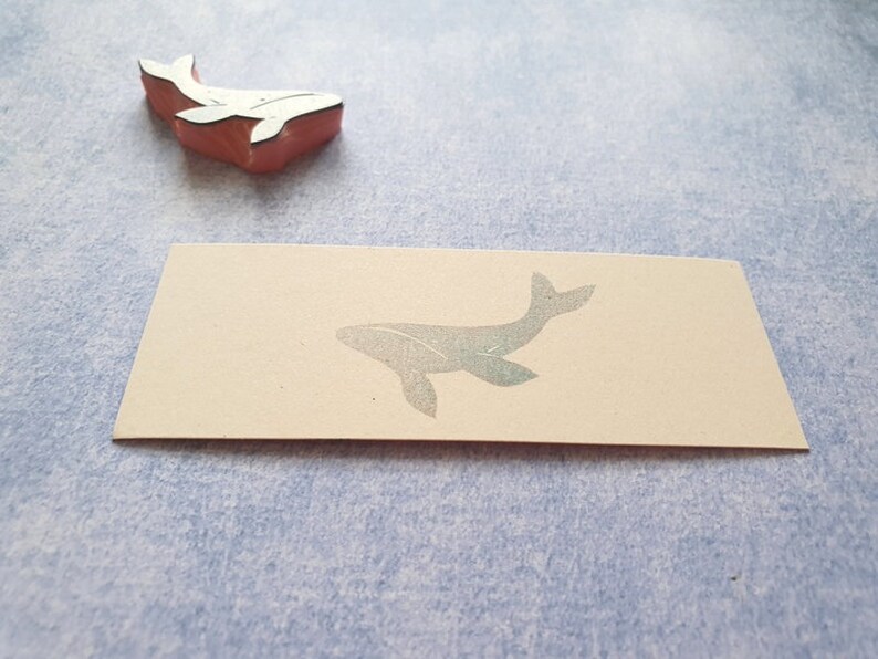 May include: A close-up of a blank card with a whale silhouette printed on it. The whale is a light gray color and is facing to the right. The card is a light beige color and has a slightly textured surface.