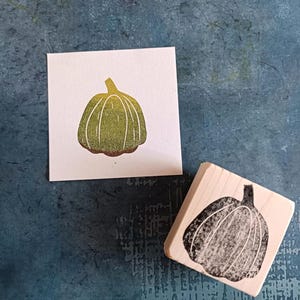 Può includere: Un timbro di zucca verde e marrone con una stampa corrispondente su una carta bianca. Il timbro è in legno e presenta un disegno di zucca nera. La stampa è un'illustrazione dettagliata di una zucca.