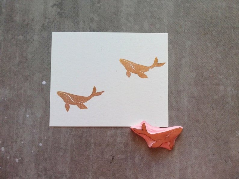May include: A white card with two brown whale silhouettes printed on it. A pink whale rubber stamp is on the table below the card.