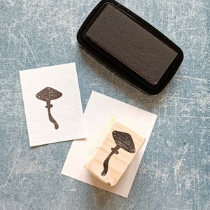 K&ouml;nnte beinhalten: Ein Stempel aus Holz mit einem Pilzmotiv, ein schwarzes Stempelkissen und zwei leere Karten. Der Stempel ist aus hellem Holz und hat ein dunkelbraunes Pilzmotiv. Das Stempelkissen ist schwarz und rechteckig. Die Karten sind wei&szlig; und rechteckig.