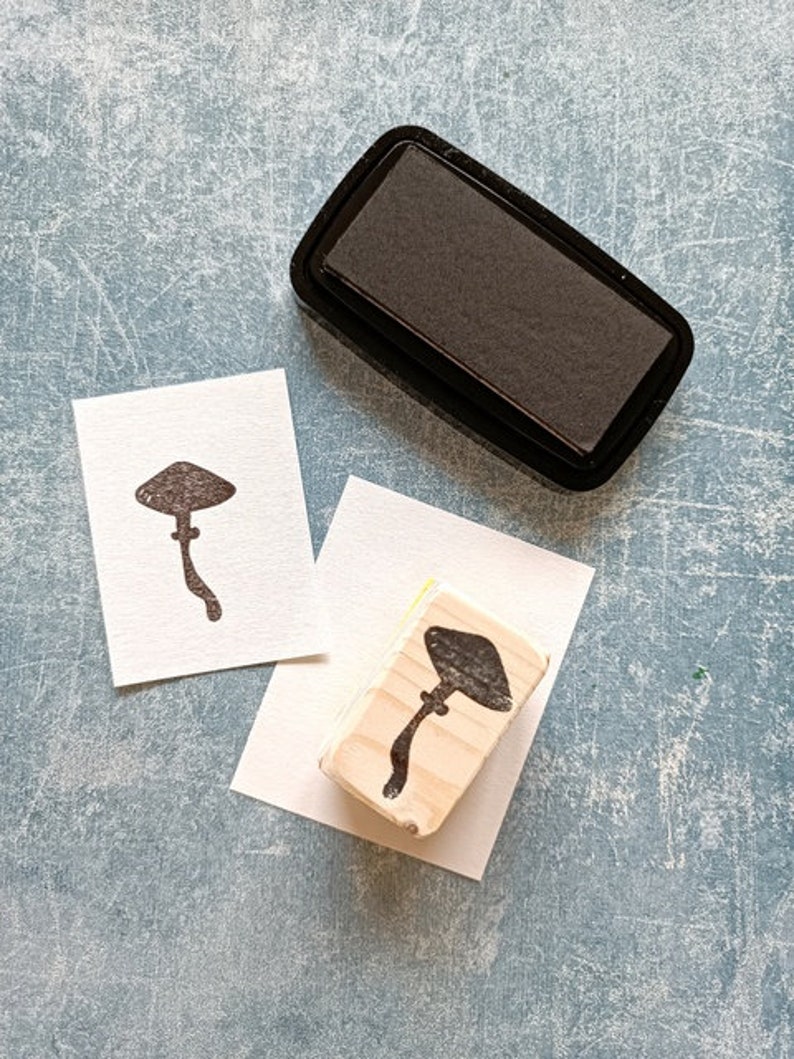 K&ouml;nnte beinhalten: Ein Stempel aus Holz mit einem Pilzmotiv, ein schwarzes Stempelkissen und zwei leere Karten. Der Stempel ist aus hellem Holz und hat ein dunkelbraunes Pilzmotiv. Das Stempelkissen ist schwarz und rechteckig. Die Karten sind wei&szlig; und rechteckig.