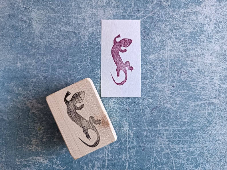 May include: A wooden stamp with a lizard design next to a stamped image of the same lizard in purple ink on a white card. The stamp is rectangular with rounded edges. The background is a textured blue surface.