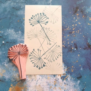 May include: A pink rubber stamp with a detailed design of a dandelion flower. The stamp is on a blue background with gold flecks. The image also shows a card with several dandelion flower designs printed in gray.