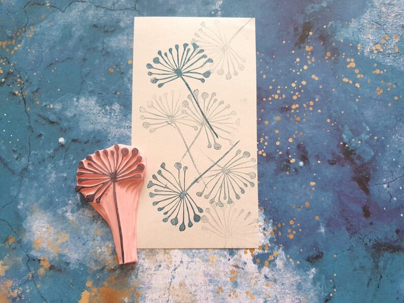May include: A pink rubber stamp with a detailed design of a dandelion flower. The stamp is on a blue background with gold flecks. The image also shows a card with several dandelion flower designs printed in gray.