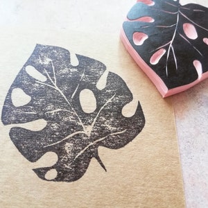 May include: A black and pink rubber stamp in the shape of a Monstera leaf, next to a leaf print on brown paper. The stamp has a pink base and a black leaf design with cut-out holes and detailed veins.