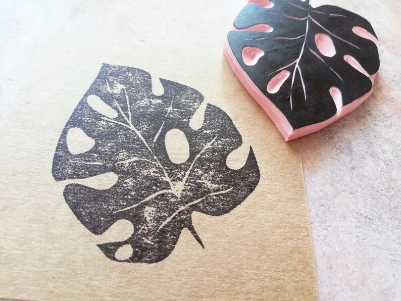 May include: A black and pink rubber stamp in the shape of a Monstera leaf, next to a leaf print on brown paper. The stamp has a pink base and a black leaf design with cut-out holes and detailed veins.