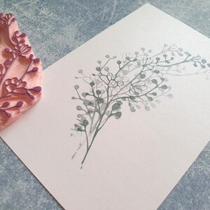 May include: A close-up of a pink rubber stamp with a floral design. The stamp has been used to create a print of a leafy branch on a white card.