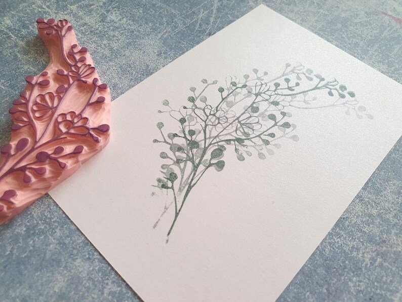 May include: A close-up of a pink rubber stamp with a floral design. The stamp has been used to create a print of a leafy branch on a white card.
