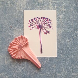 May include: A pink rubber stamp with a detailed design of a flower with a long stem. The stamp is used to create a print of a flower with purple and pink petals.