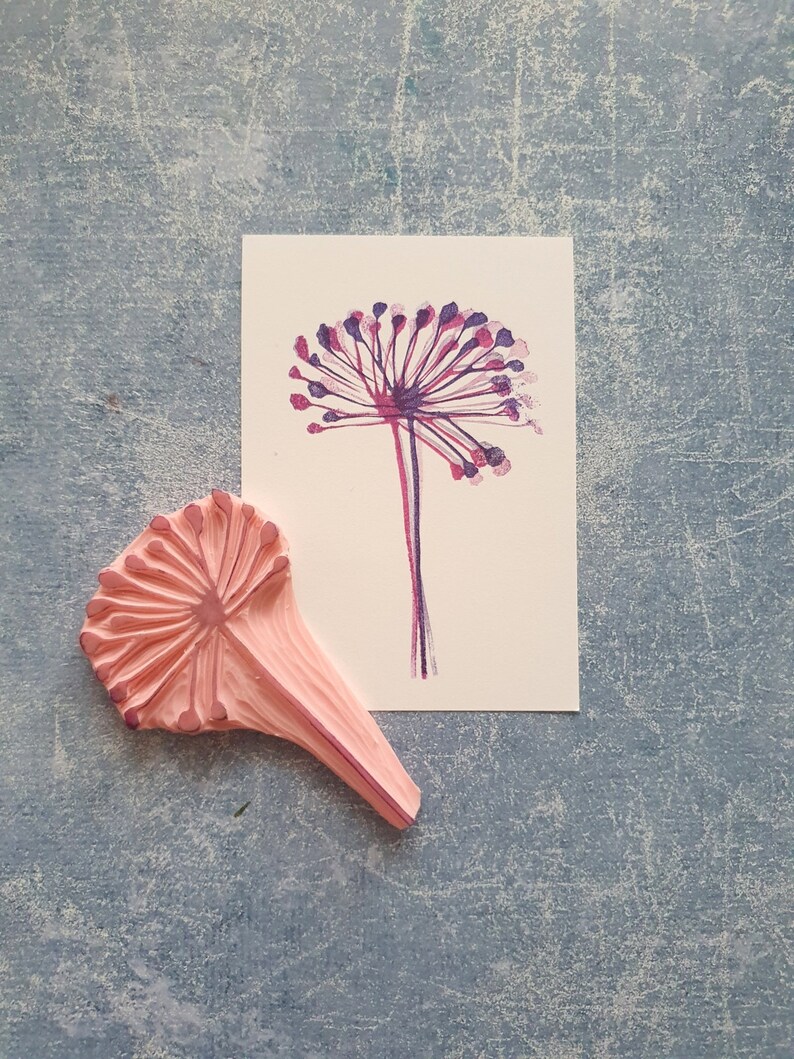 May include: A pink rubber stamp with a detailed design of a flower with a long stem. The stamp is used to create a print of a flower with purple and pink petals.