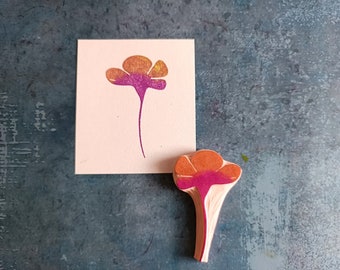 May include: A wooden stamp with a pink and orange flower design. The stamp is used to create a print of the flower on paper.