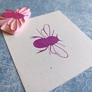 May include: A pink rubber stamp with a flower-like design next to a stamped image of a purple insect on a white card. The stamp has a layered, textured appearance. The insect image has a simple, stylized outline.
