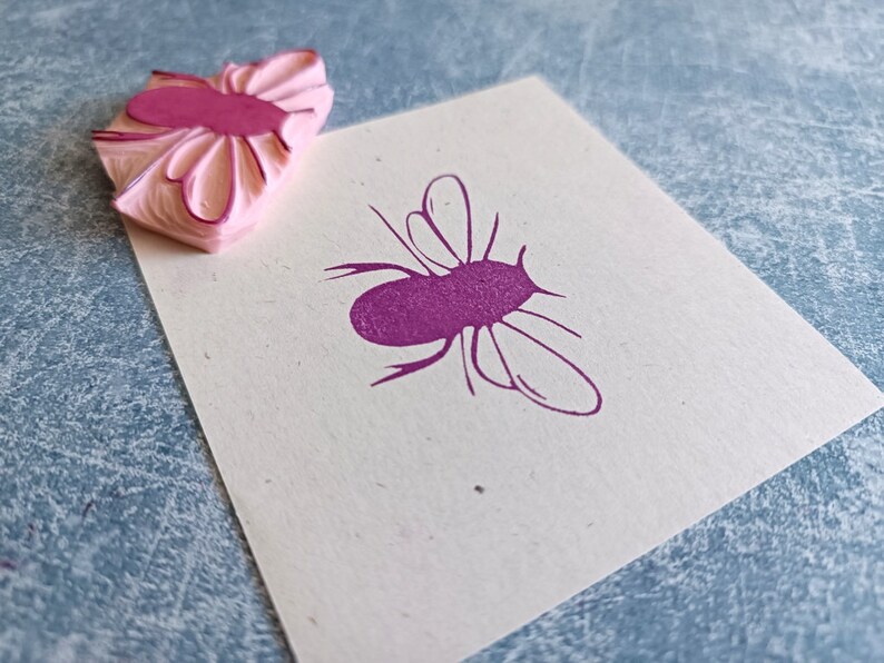 May include: A pink rubber stamp with a flower-like design next to a stamped image of a purple insect on a white card. The stamp has a layered, textured appearance. The insect image has a simple, stylized outline.