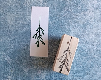 May include: A wooden stamp with a green leafy branch design. The stamp is on a blue background and a printed image of the design is next to it.