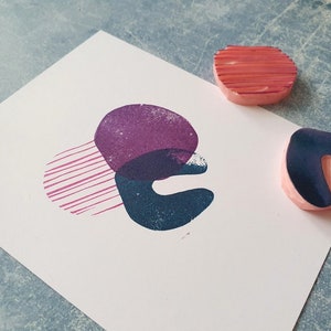 May include: An abstract art print featuring overlapping shapes in purple, teal, and pink. Two wooden stamps with pink bases and colored tops are next to the print. One stamp is a curved shape, and the other is a rounded shape with horizontal lines.