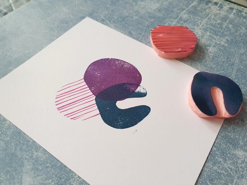 May include: An abstract art print featuring overlapping shapes in purple, teal, and pink. Two wooden stamps with pink bases and colored tops are next to the print. One stamp is a curved shape, and the other is a rounded shape with horizontal lines.